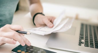 Ein Unternehmer sitzt an seinem Schreibtisch und rechnet mit Stift, Taschenrechner und Papier seine Mehrwertsteuersenkung aus.
