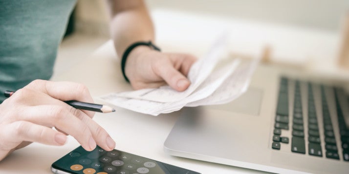 Ein Unternehmer sitzt an seinem Schreibtisch und rechnet mit Stift, Taschenrechner und Papier seine Mehrwertsteuersenkung aus.