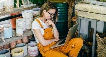 Eine Frau in Arbeitskleidung sitzt in ihrer Werkstatt und netzwerkt dabei lässig auf ihrem Laptop.
