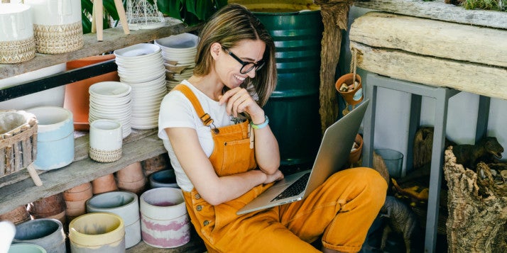 Eine Frau in Arbeitskleidung sitzt in ihrer Werkstatt und netzwerkt dabei lässig auf ihrem Laptop.