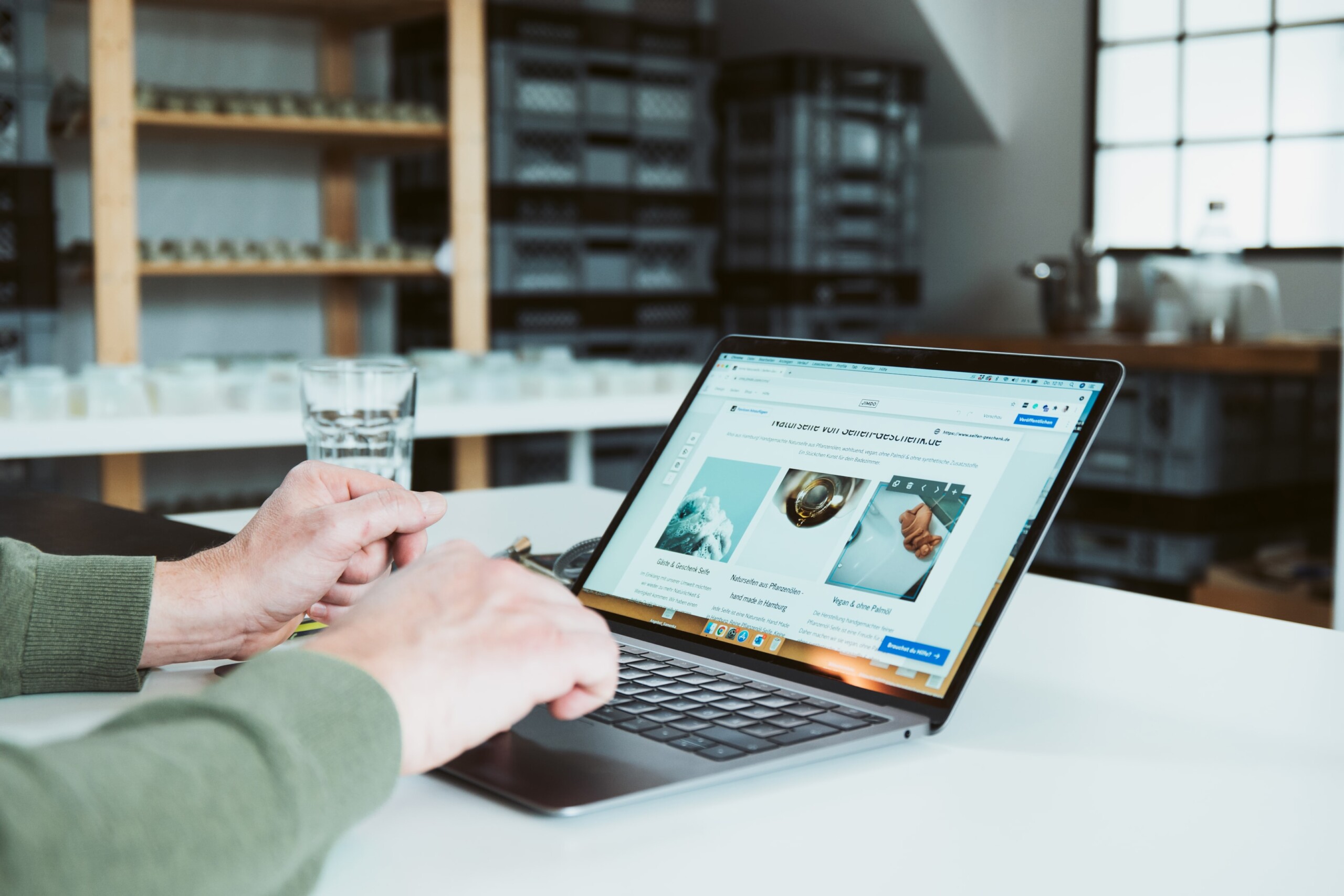 Person arbeitet am Laptop an einer Jimdo-Website. Im Hintergrund stehen Regale mit Gläsern und Kisten in einem hellen Atelier oder Büro.
